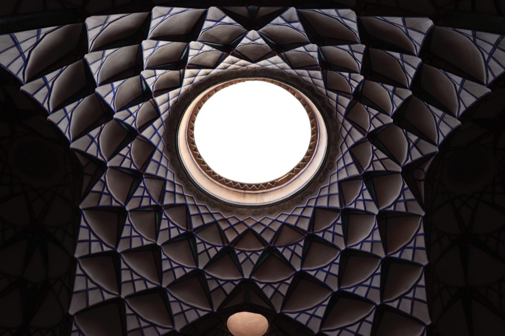 Looking up at an ornate tiled dome with a bright circular opening at its center