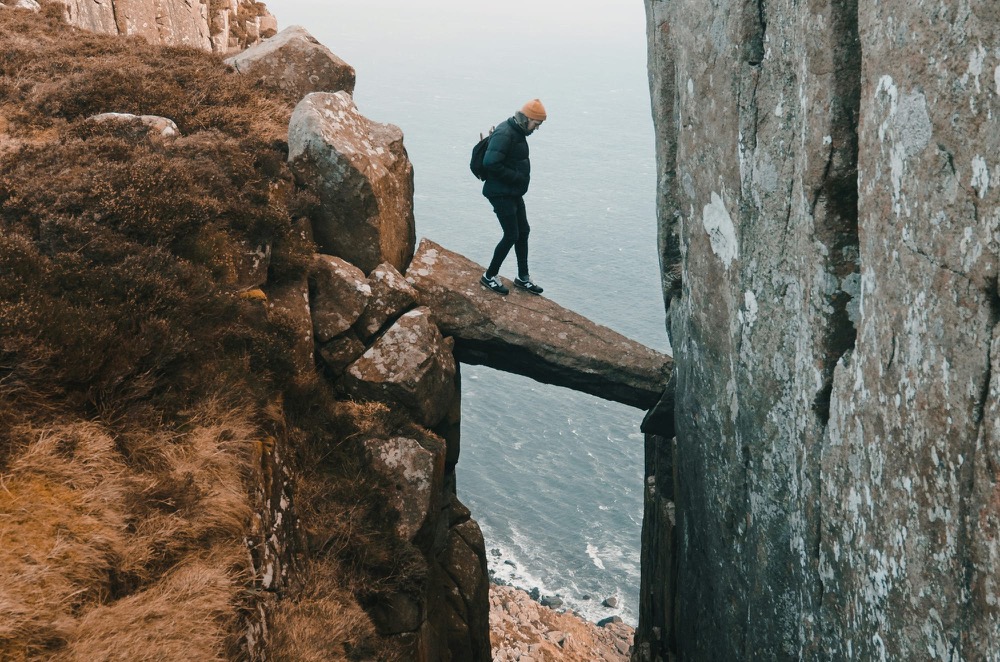 A hiker crossing a narrow rock arch between two cliffs above the sea