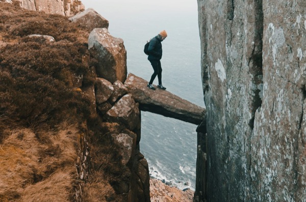 A hiker crossing a narrow rock arch between two cliffs above the sea