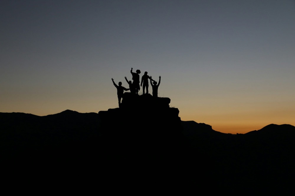 Silhouettes of a small group standing on a rocky summit with their arms raised at dusk