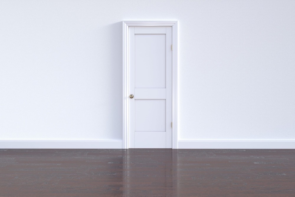 A single closed white door in an empty white room with a dark wood floor