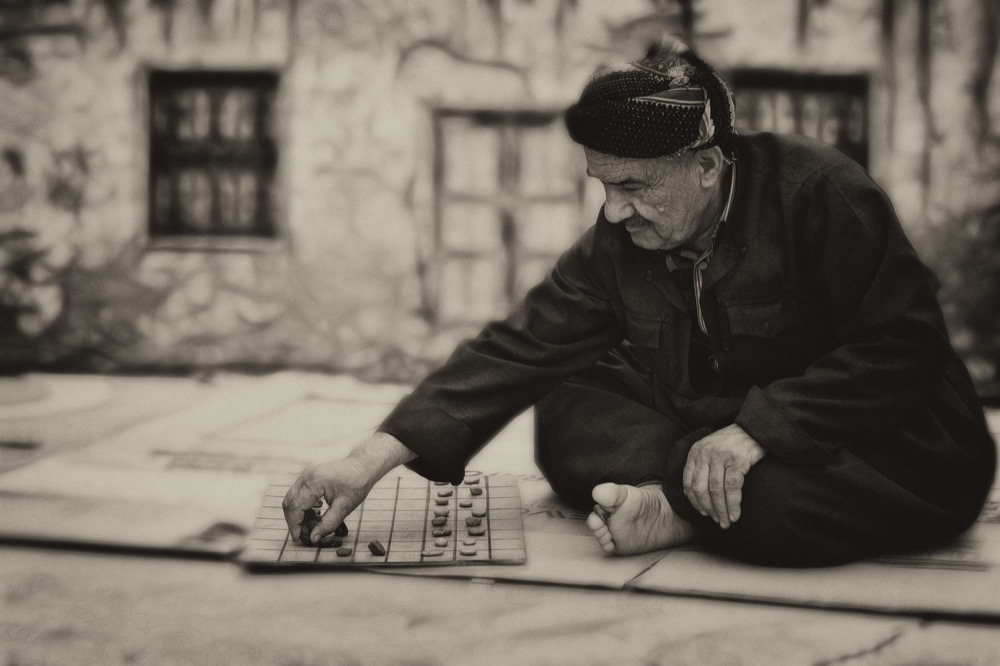An older man crouched over a board game laid out on the ground outdoors
