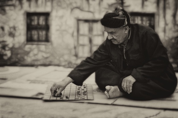 An older man crouched over a board game laid out on the ground outdoors