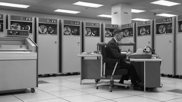 A vintage photograph of a man working at a desk in a room of mainframe computers